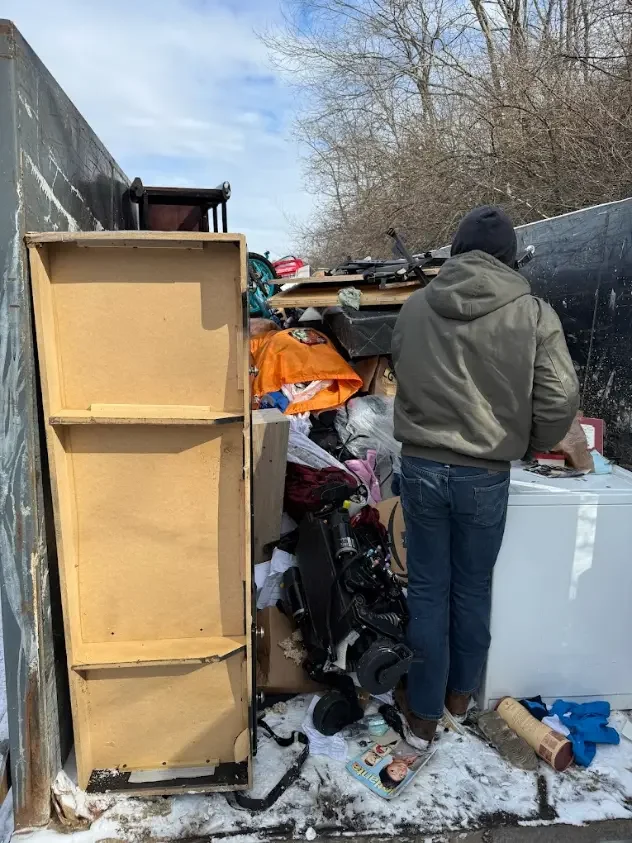 Person in gray hoodie and jeans sorting through clutter outside, with a bookshelf, camera gear, and trash around, near a black truck and leafless trees on a cold day.