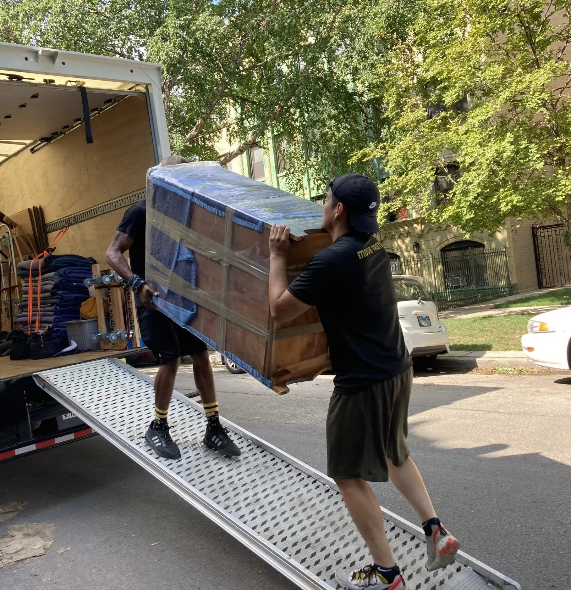 Two men loading furniture onto a truck, one inside the truck and the other on a ramp outside. The man outside is carrying a large wooden item wrapped in plastic. They are outside on a residential street with trees and parked cars.