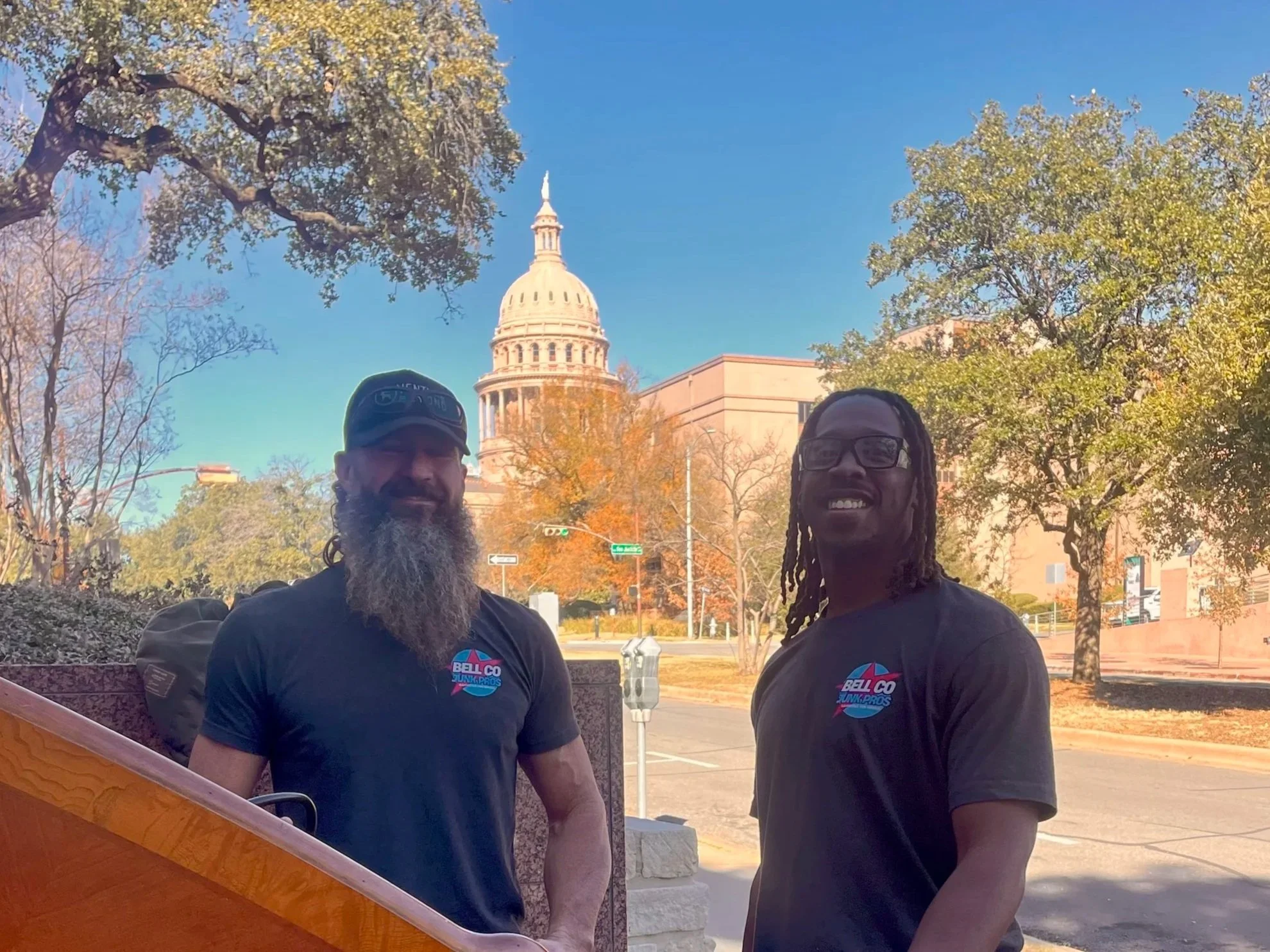 Two men pose outdoors in front of the United States Capitol building, surrounded by trees with autumn foliage. Both are smiling and wearing black T-shirts with a colorful logo that reads 'BELL CO' and 'JUNAPROS'.