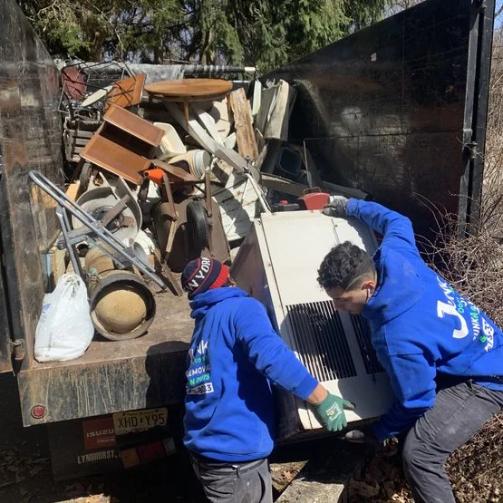 Two men wearing blue jackets are unloading items from a truck filled with furniture and household items, including chairs, a lamp, and a large black container.