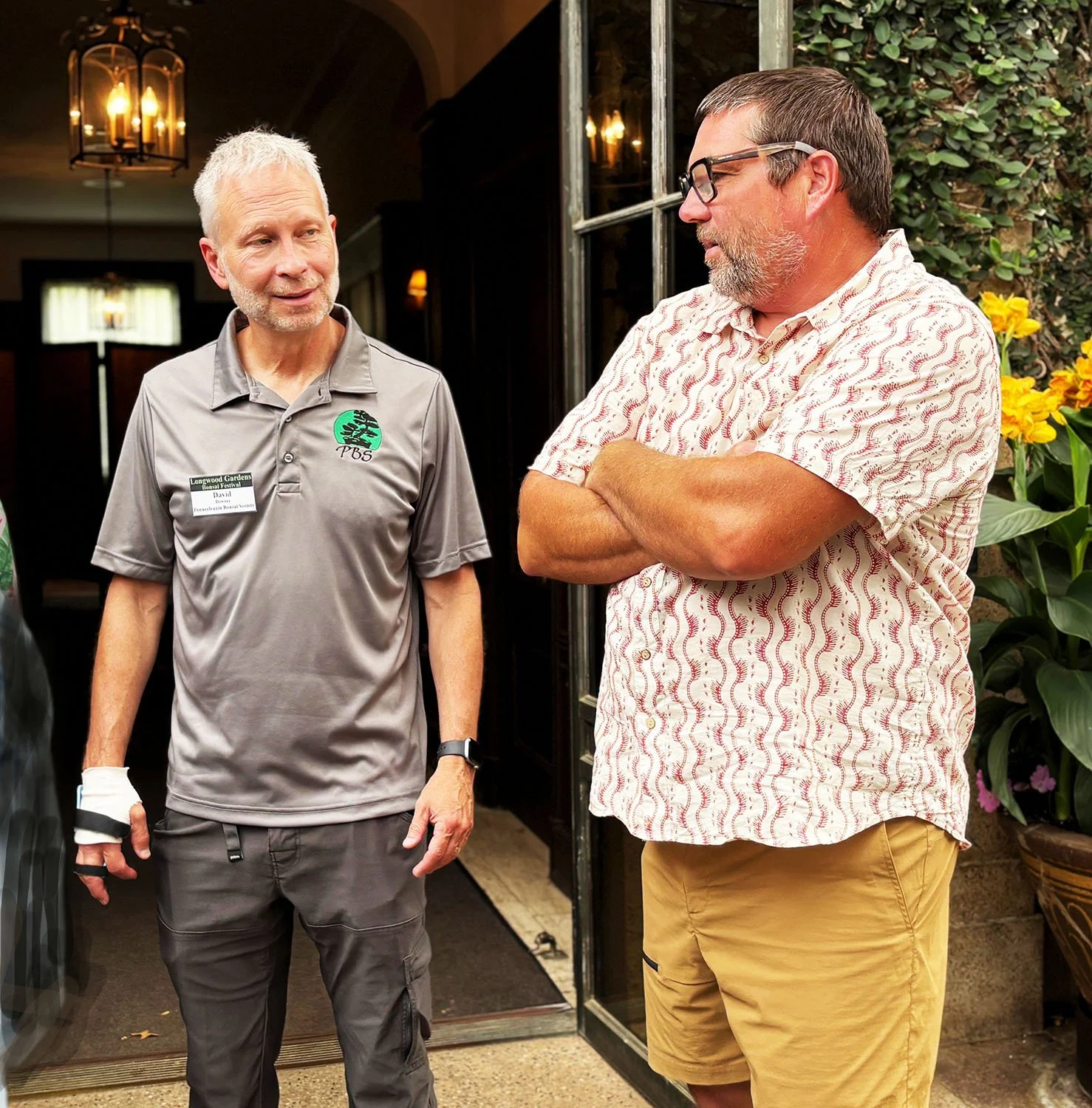 Two men standing and engaging in conversation outside a restaurant or cafe, with one wearing a gray polo shirt with a logo and the other in a patterned short-sleeve shirt, surrounded by potted plants.
