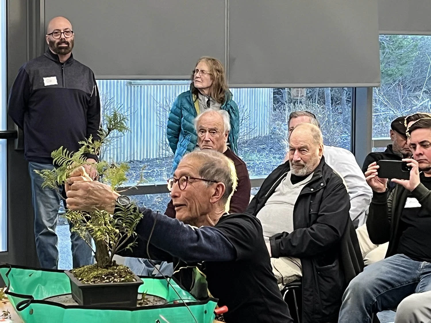 A group of people attending a bonsai workshop or presentation, with a man holding a small bonsai tree and some participants taking photos in a room with large windows and winter scenery outside.