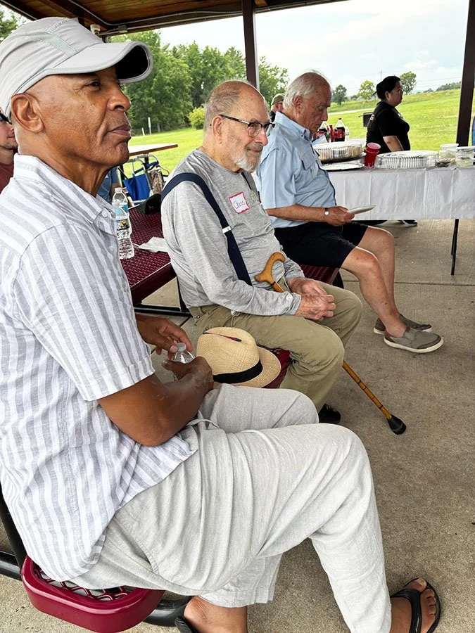Four men sitting in a row under a pavilion at an outdoor event, with a table of food and drinks behind them and a grassy field in the background.