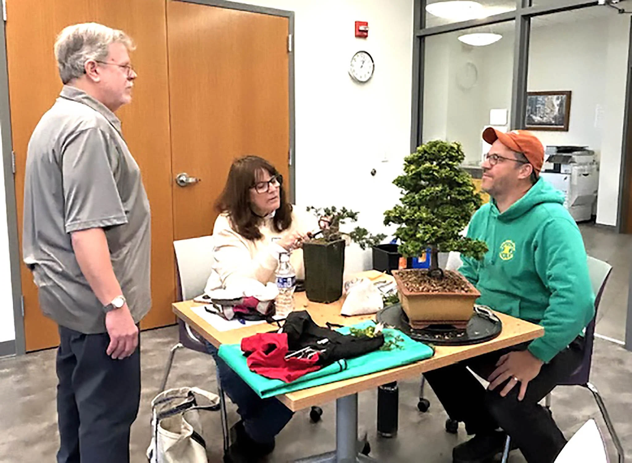Three people are gathered around a table indoors with plants on display, with one man standing and two seated. The scene is in a well-lit room with glass walls and a clock on the wall.