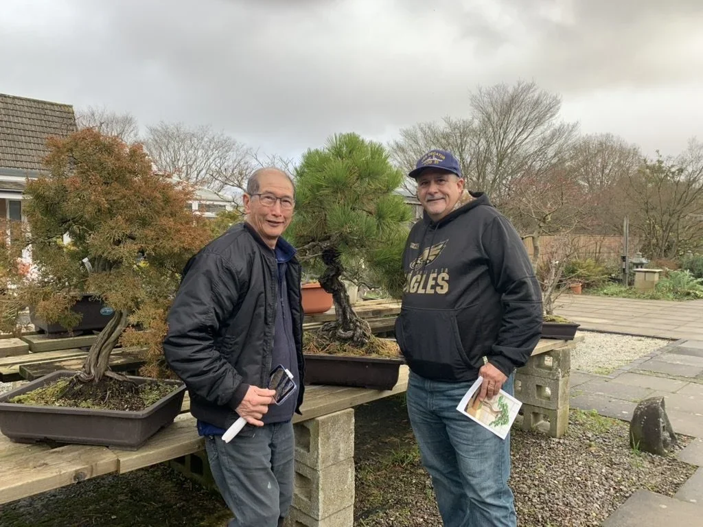 Two men standing outdoors near bonsai trees on wooden tables, holding papers, with leafless trees and overcast sky in the background.