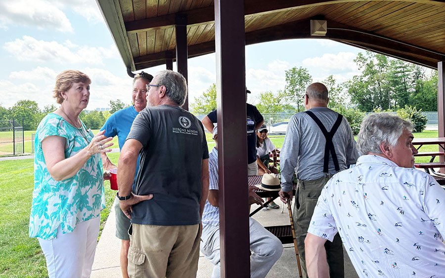 Group of people talking and sitting outdoors under a pavilion on a bright, partly cloudy day.