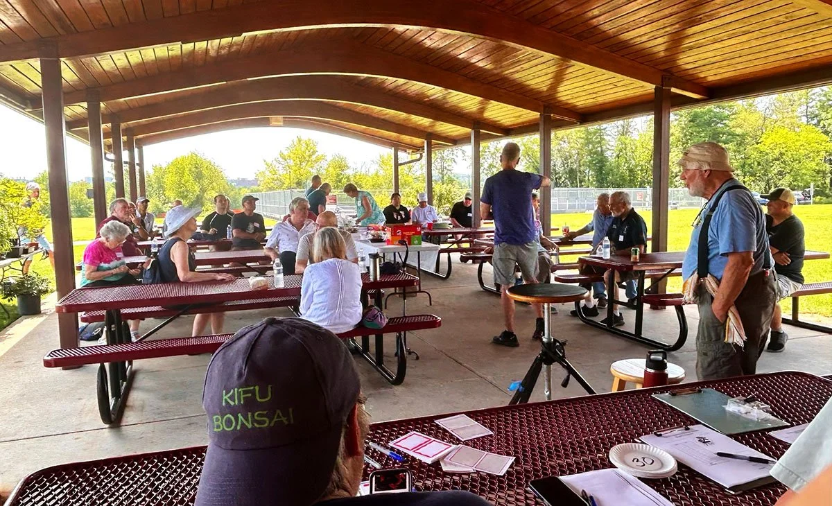 People gathered in an outdoor pavilion with a wooden roof, sitting at picnic tables, listening to a speaker in casual attire, surrounded by trees and a grassy field.