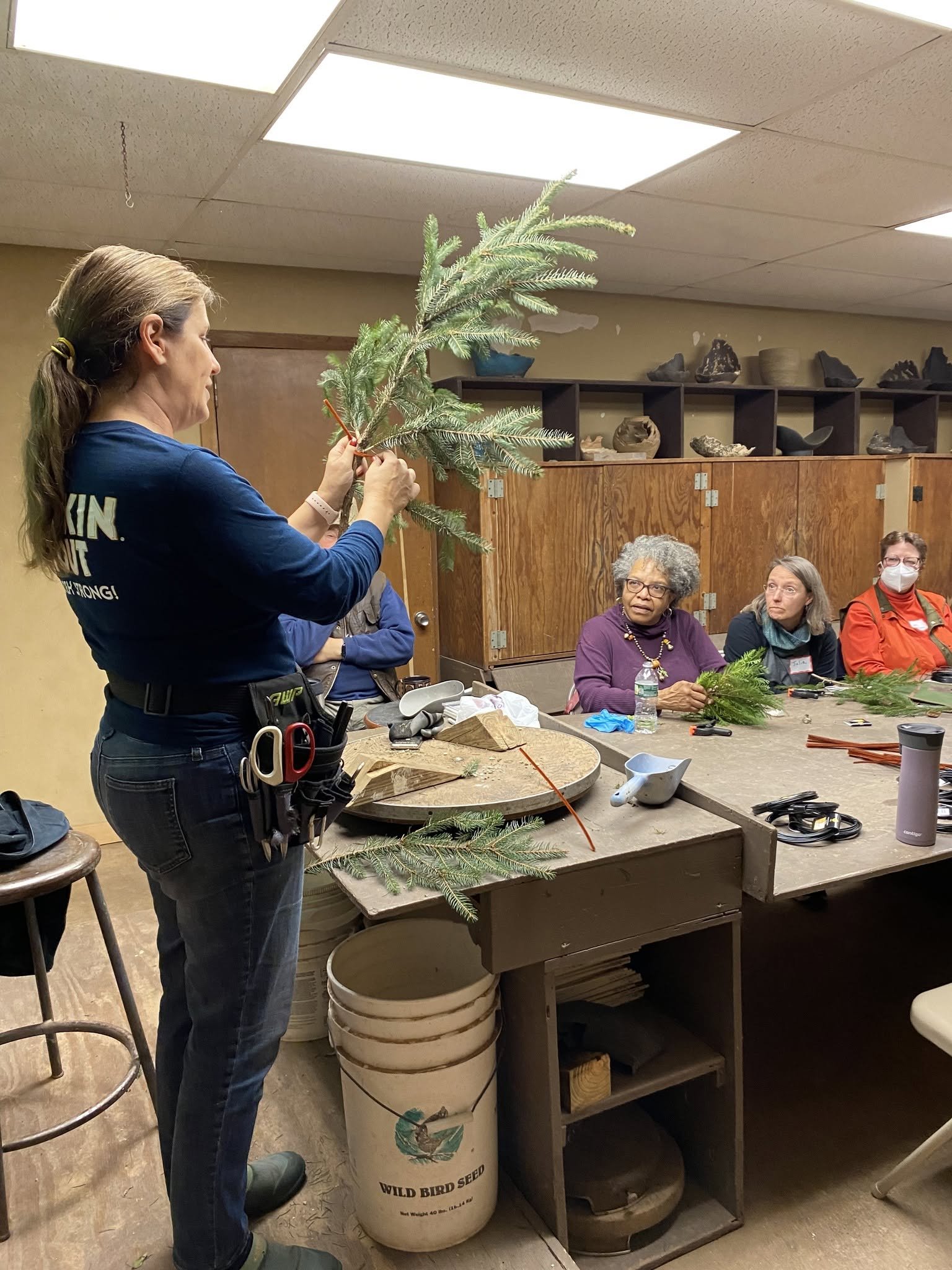 A woman with a ponytail is standing at a workbench, holding a branch of an evergreen tree. Several people sitting at the table are observing her, with some wearing masks. The workspace has various tools, a large white bucket labeled 'Wild Bird Seed' 