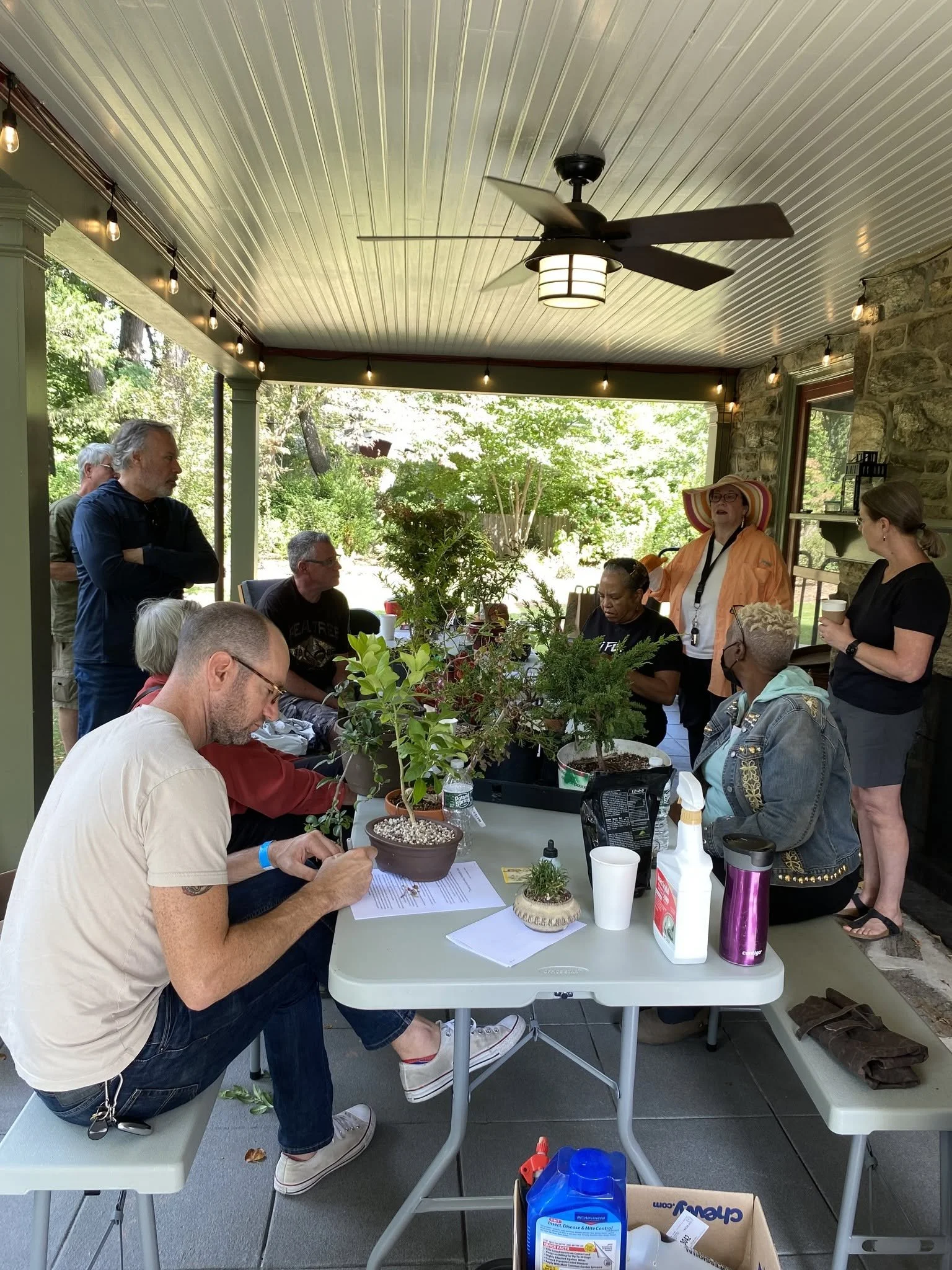 People gathered around a table with plants, engaged in conversation, inside a porch area with green foliage visible outside.