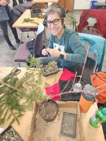 A woman with glasses and a watch sitting at a table, smiling, in a classroom or workshop setting. The table has various potted plants, gardening tools, and a water bottle. She is wearing a teal shirt with a star on it, and there are other people and 