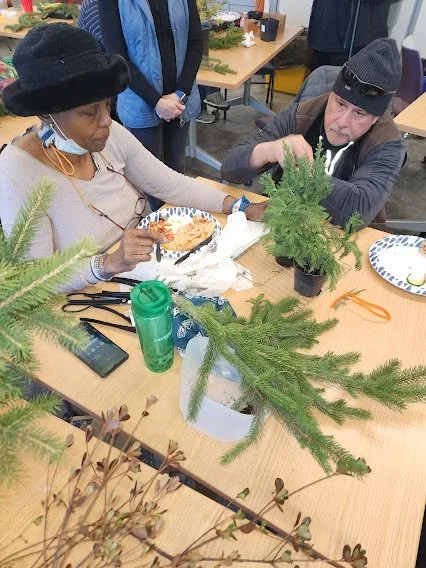 Two people sitting at a table working on a craft project with evergreen branches, including a small potted pine tree. Both are wearing winter hats.