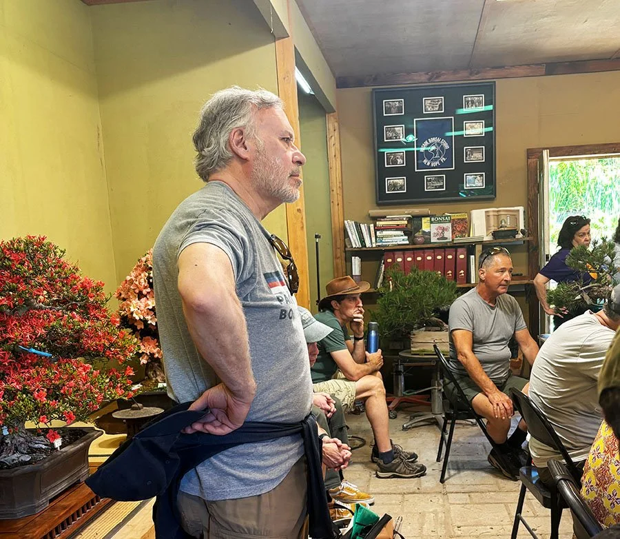 A group of people inside a room, some sitting and some standing, with a man in the foreground listening attentively. The room has plants, books, and a framed display on the wall.