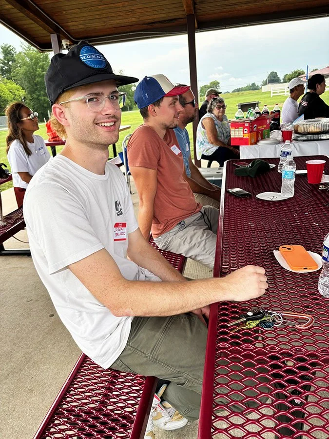 A group of people sitting at a covered outdoor picnic table during daytime, with food, drinks, and snacks on the table, and a grassy field in the background.