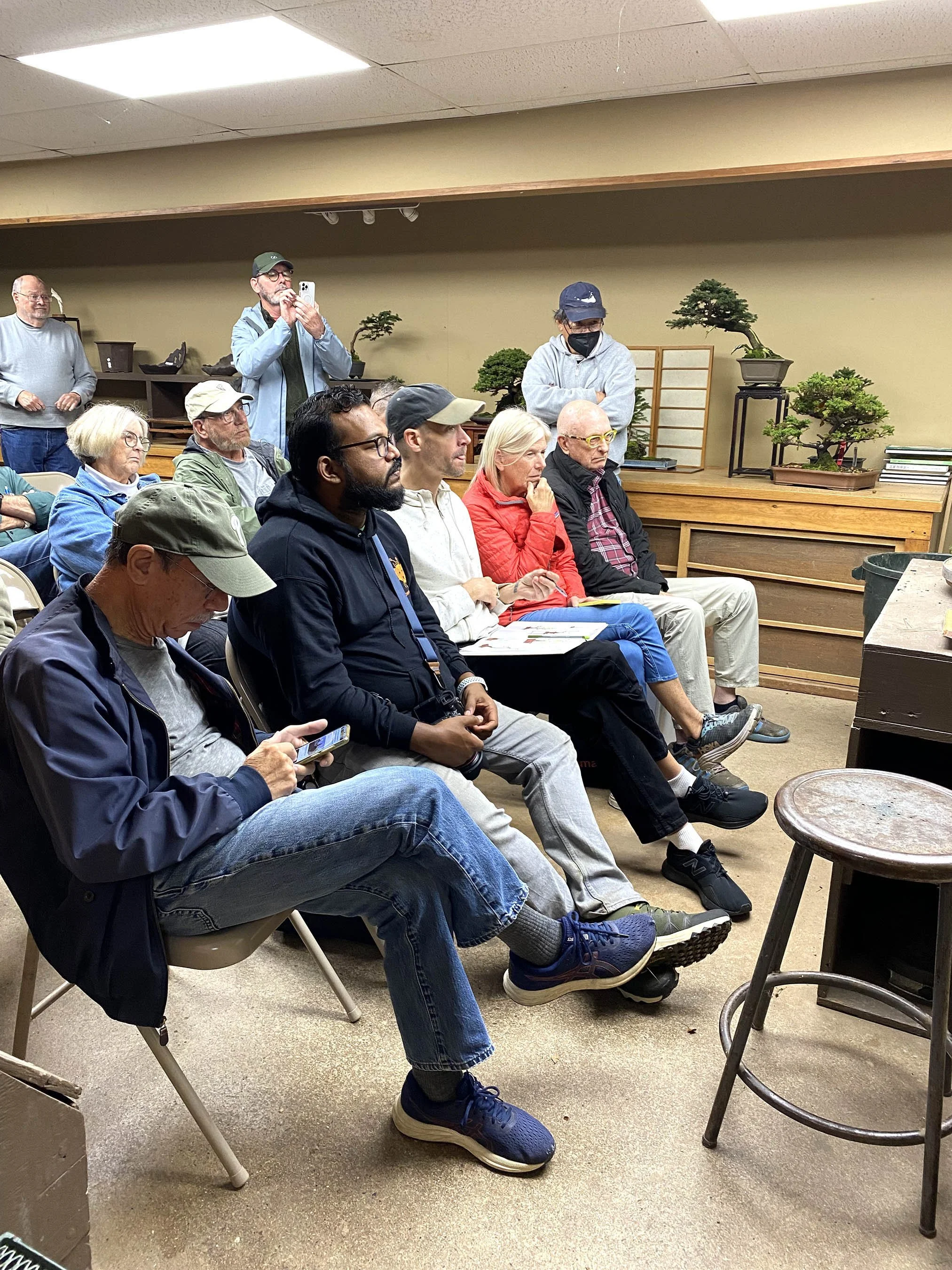 A group of people seated in chairs in a room, listening to a presentation or lecture. Two people are standing at the back, one taking a photo with a phone and the other wearing a mask and looking toward the front. The room has a wooden shelf with bon