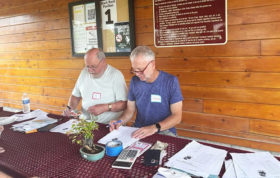 Two men sitting at a table, working with papers, pens, and a calculator, with a small bonsai tree in a pot in front of them and a water bottle on the side. The background features wood-paneled walls and signs.