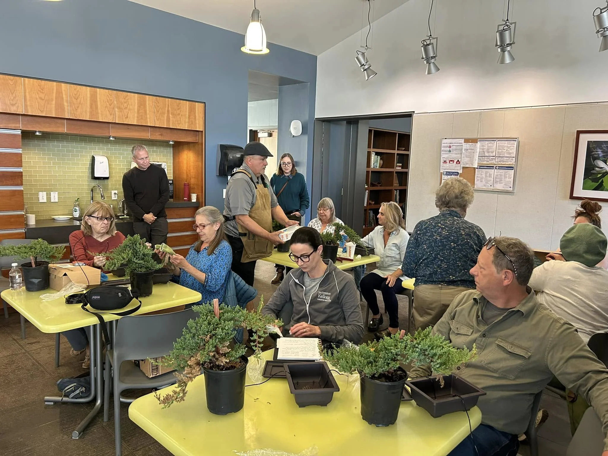 People gathered in a room with tables covered in small potted plants, engaging in a plant workshop or class.