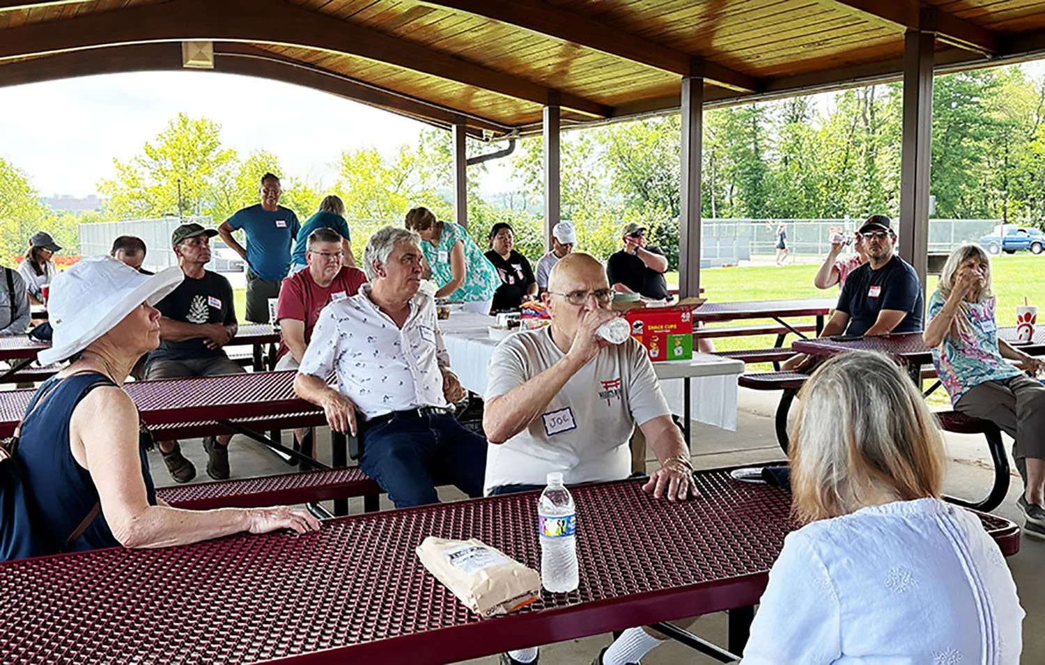 People seated at picnic tables under a pavilion during an outdoor gathering, with some drinking water and others socializing, green trees and a field in the background.
