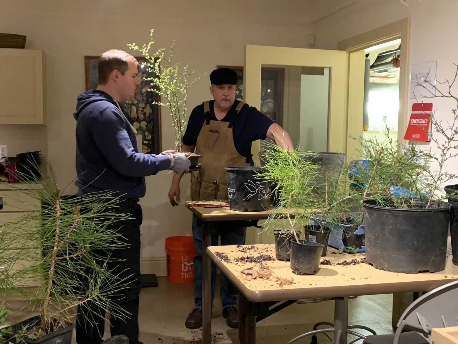 Two men working with plants in an indoor setting. One man is wearing a dark hoodie and gloves, and the other is wearing a dark shirt with an apron and a black cap. They are standing at tables with potted plants and gardening supplies.