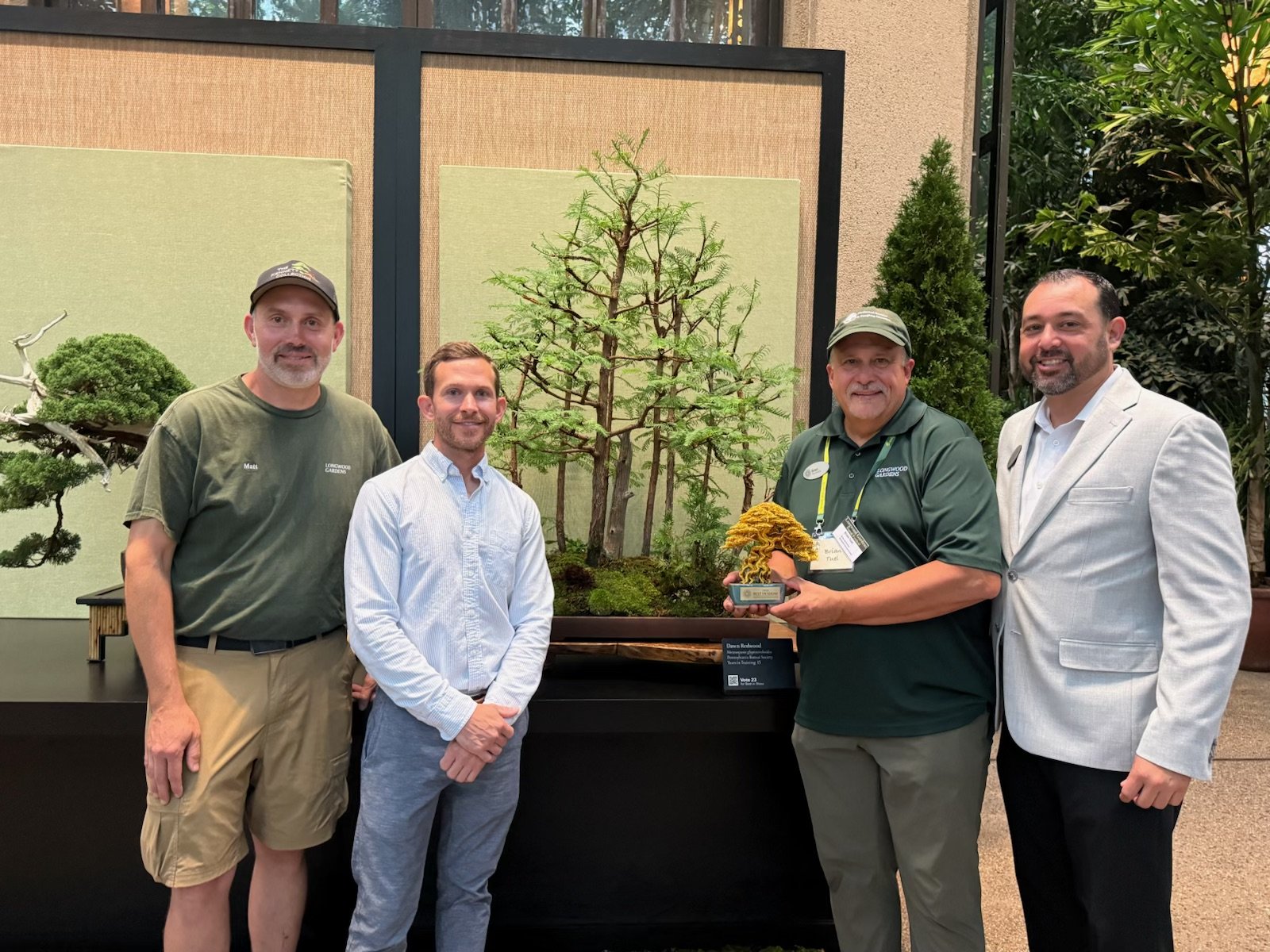 Four men standing together indoors in front of a bonsai display, with one holding a Bonsai tree award, smiling for the photo.