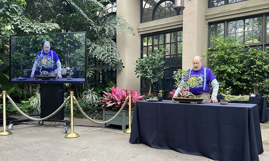 A man working on a floral arrangement at a table inside a greenhouse or conservatory with large windows, plants, and a television monitor showing his image.