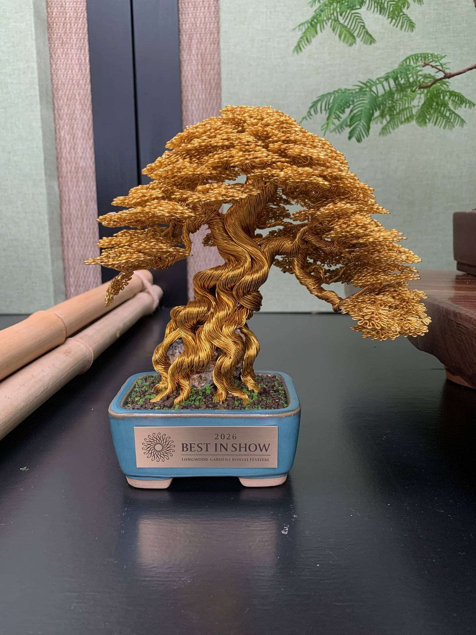A metallic gold-colored wire bonsai tree sculpture in a blue pot, labeled as 'Best in Show' at the 2026 Longwood Gardens Bonsai Festival, with bamboo and greenery in the background.