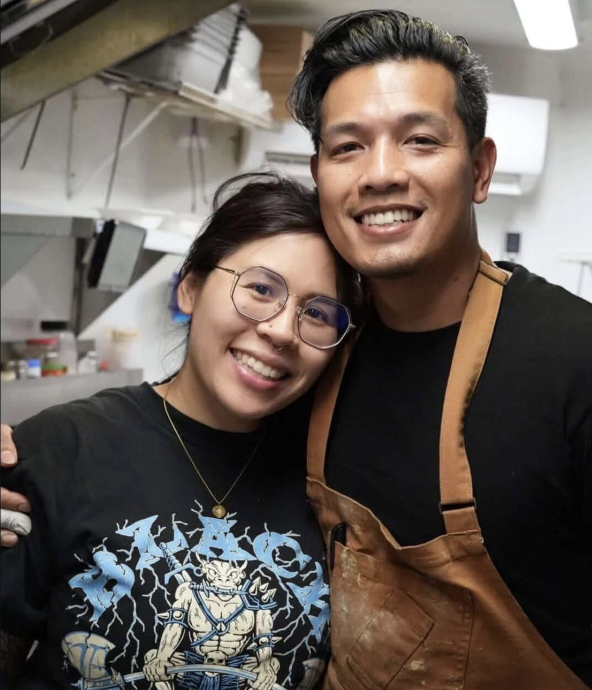 A smiling woman with glasses and a necklace, and a smiling man wearing an apron, taking a selfie together in a kitchen.