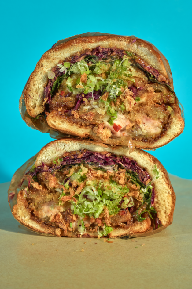 Close-up of a halved fried chicken sandwich with lettuce, pickles, and sauce, on a bun, against a bright blue background.