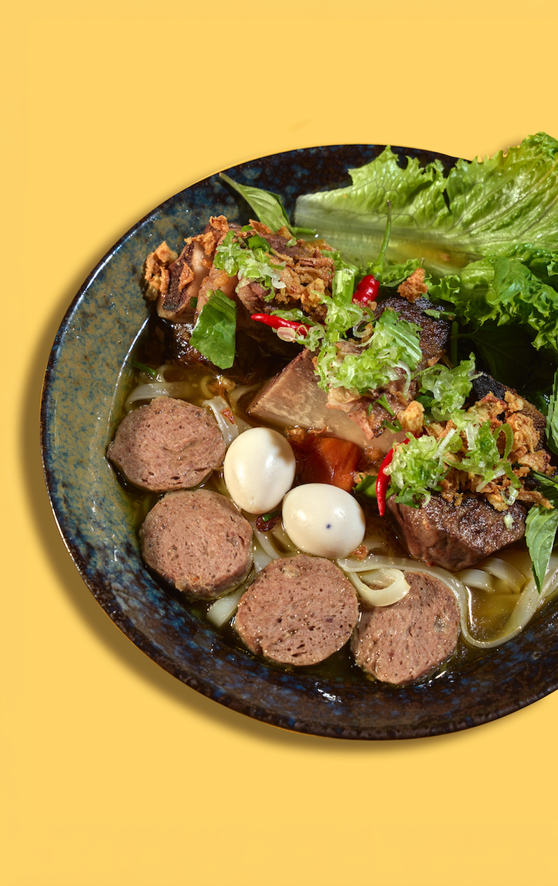 Bowl of noodle soup with meatballs, beef, quail eggs, vegetables, and herbs.