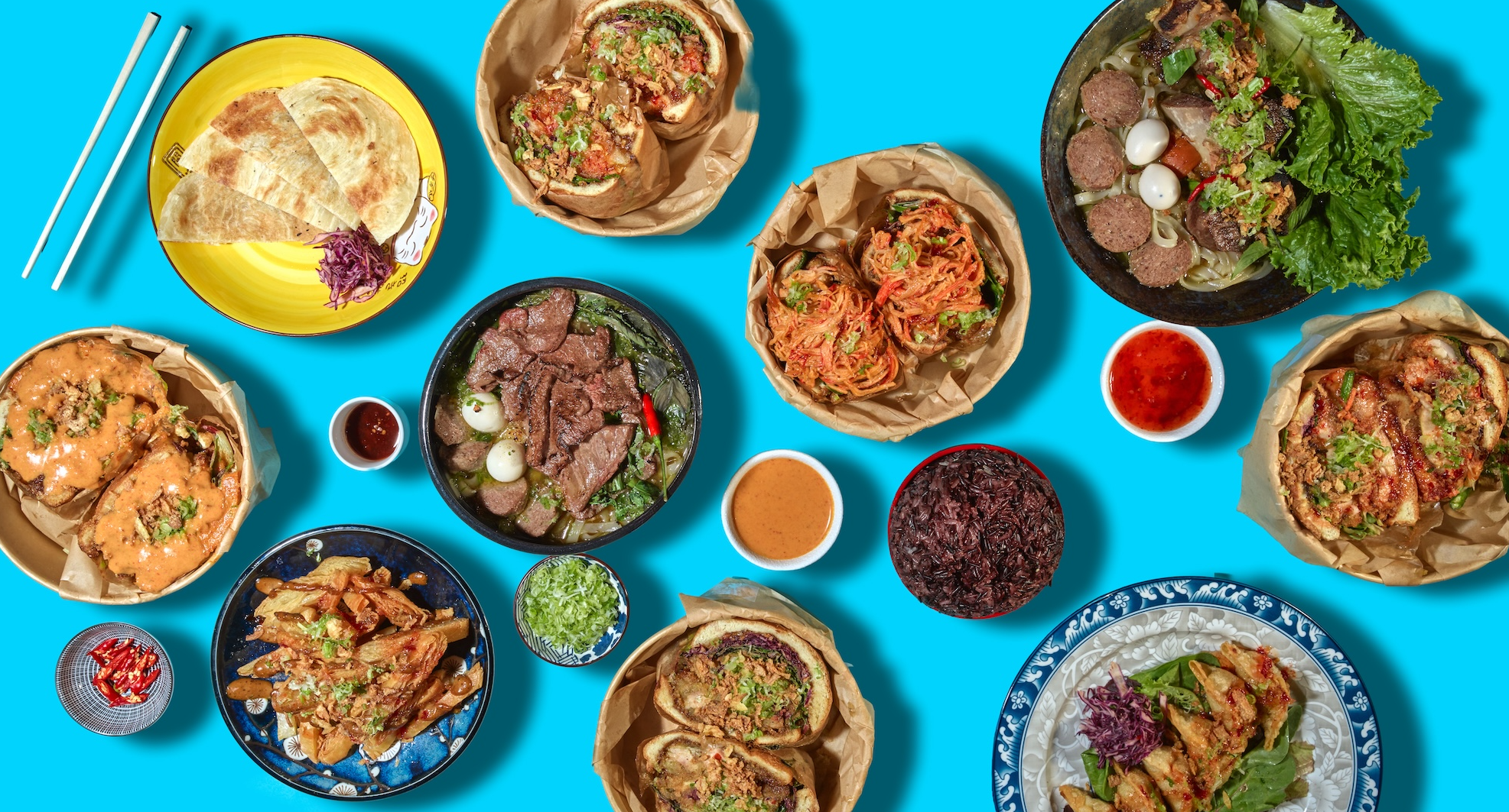 A variety of Asian dishes including wraps, noodles, grilled meats, rice, and small bowls of sauces, all arranged on a bright blue surface.