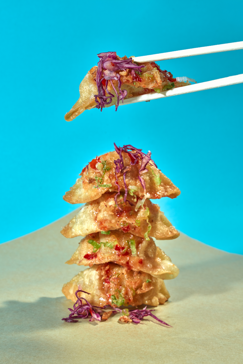 Stacked fried dumplings topped with chopped green onions and purple cabbage shreds, with a chopstick holding one dumpling against a blue background.