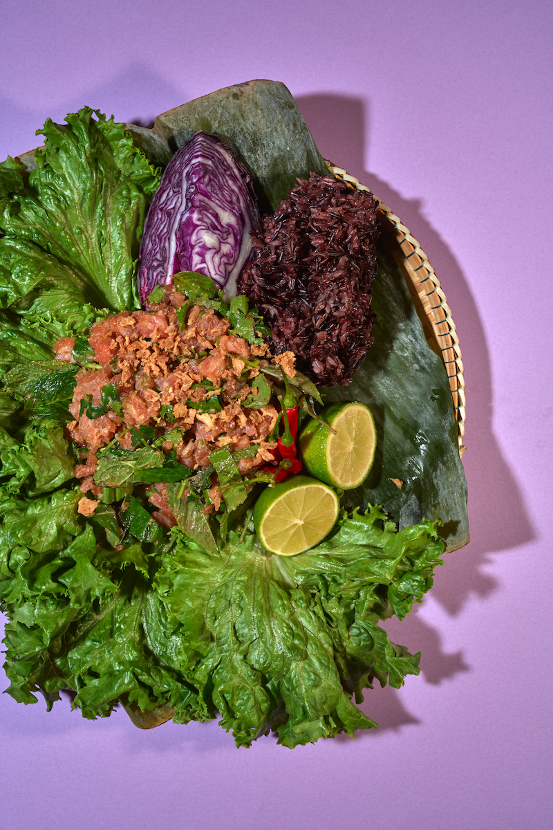 A basket of Asian food with lettuce, purple cabbage, cooked rice, lime slices, and a meat and vegetable mixture on a banana leaf.