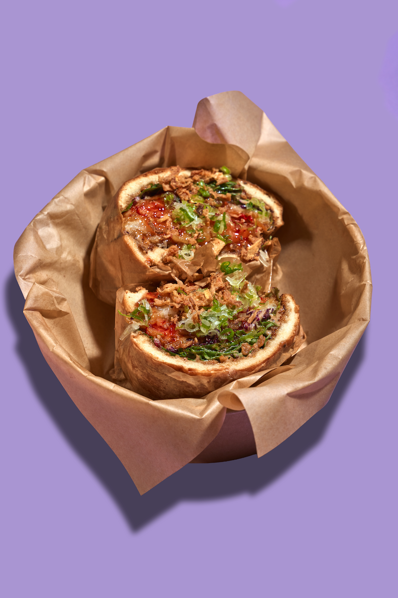 A close-up of a steamed bun sliced in half, filled with ground meat, chopped green onions, and sauce, served in brown paper on a purple background.