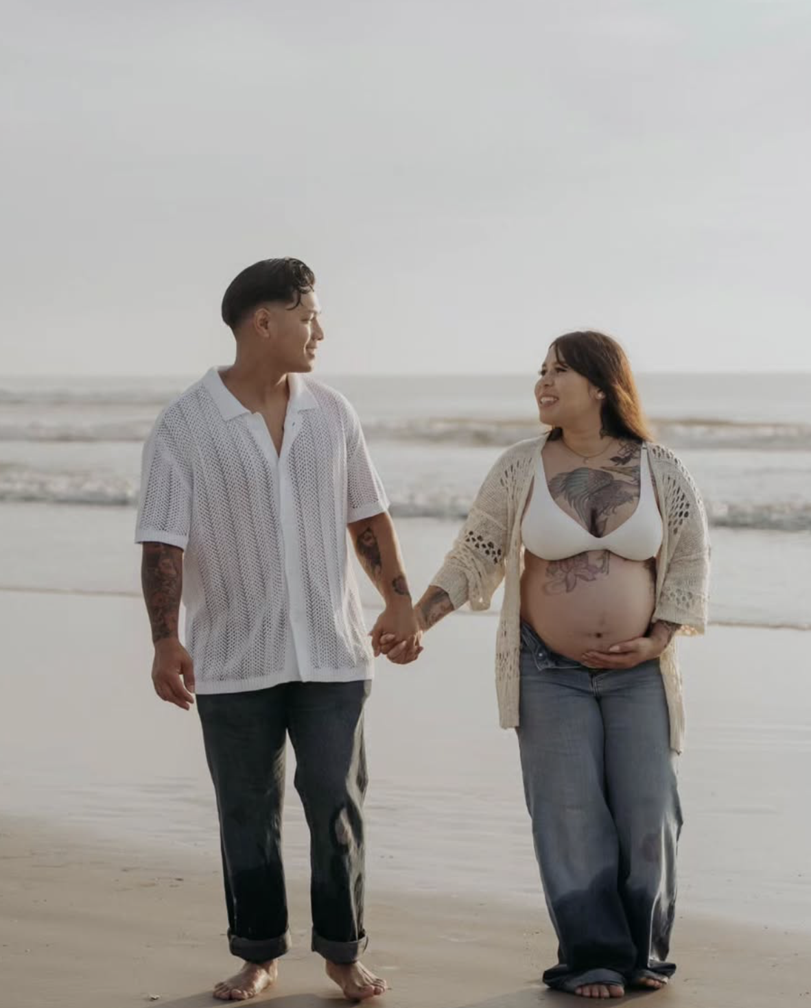 A couple walking hand in hand on the beach, smiling at each other. The woman is pregnant and has tattoos on her chest and stomach, and the man has tattoos on his arms. They are dressed casually, with beach attire, and the ocean is in the background.