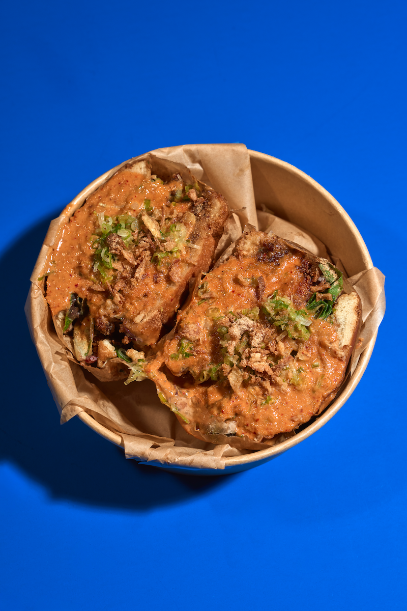 Two fried sandwiches topped with sauce, chopped green onions, and crispy onions in a brown paper-lined container against a blue background.