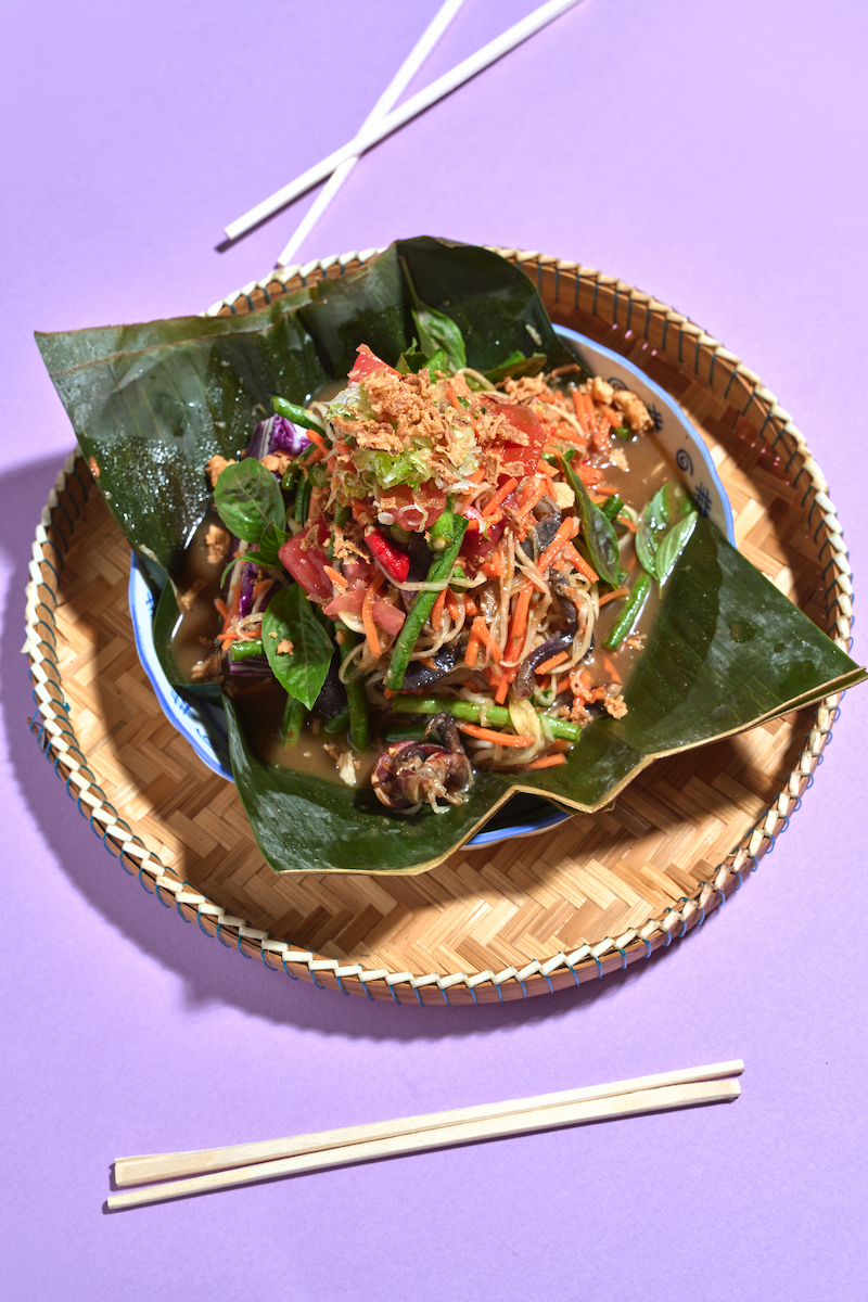 A plate of Asian vegetable stir-fry with noodles, served on banana leaves, placed in a woven basket on a purple background with chopsticks.