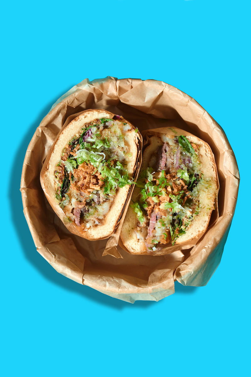 Close-up of a halved stuffed bread roll filled with minced meat, vegetables, and sauce, placed in a brown paper-lined bowl against a purple background.