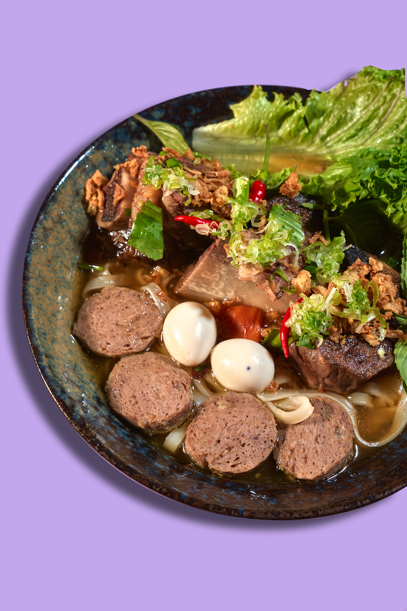 A bowl of Vietnamese pho with beef meatballs, slices of grilled beef, quail eggs, fresh greens, and herbs in a brothy soup.