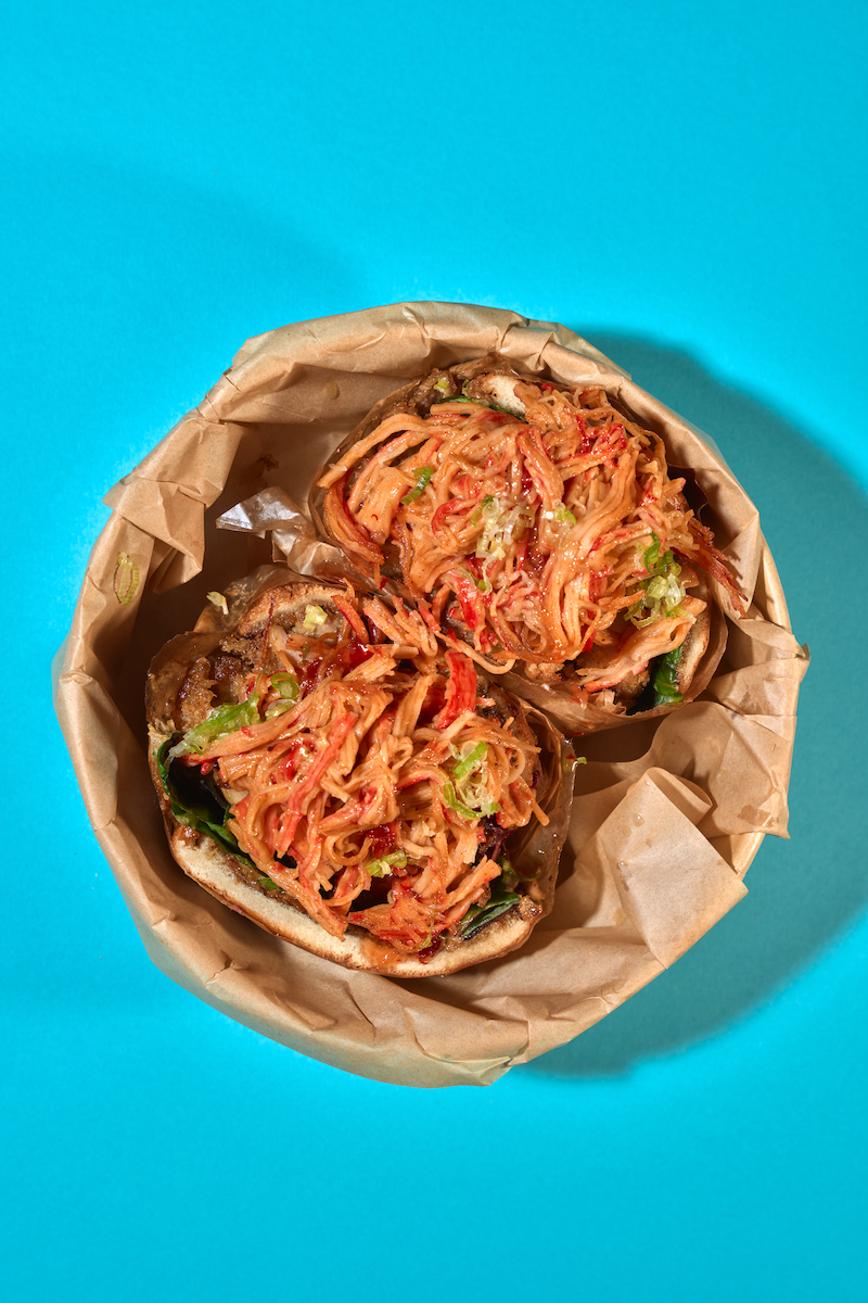 Two steamed buns filled with shredded spicy chicken and vegetables, wrapped in brown paper, against a bright blue background.