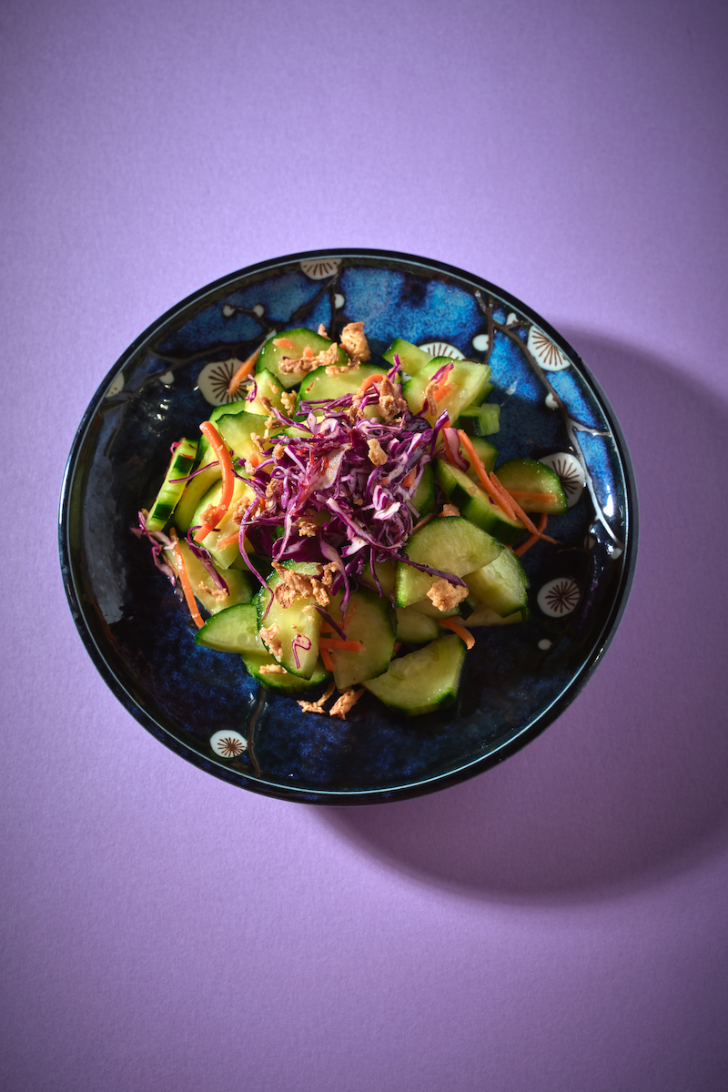A bowl of cucumber salad with shredded purple cabbage, shredded carrots, and crushed nuts, on a purple surface.