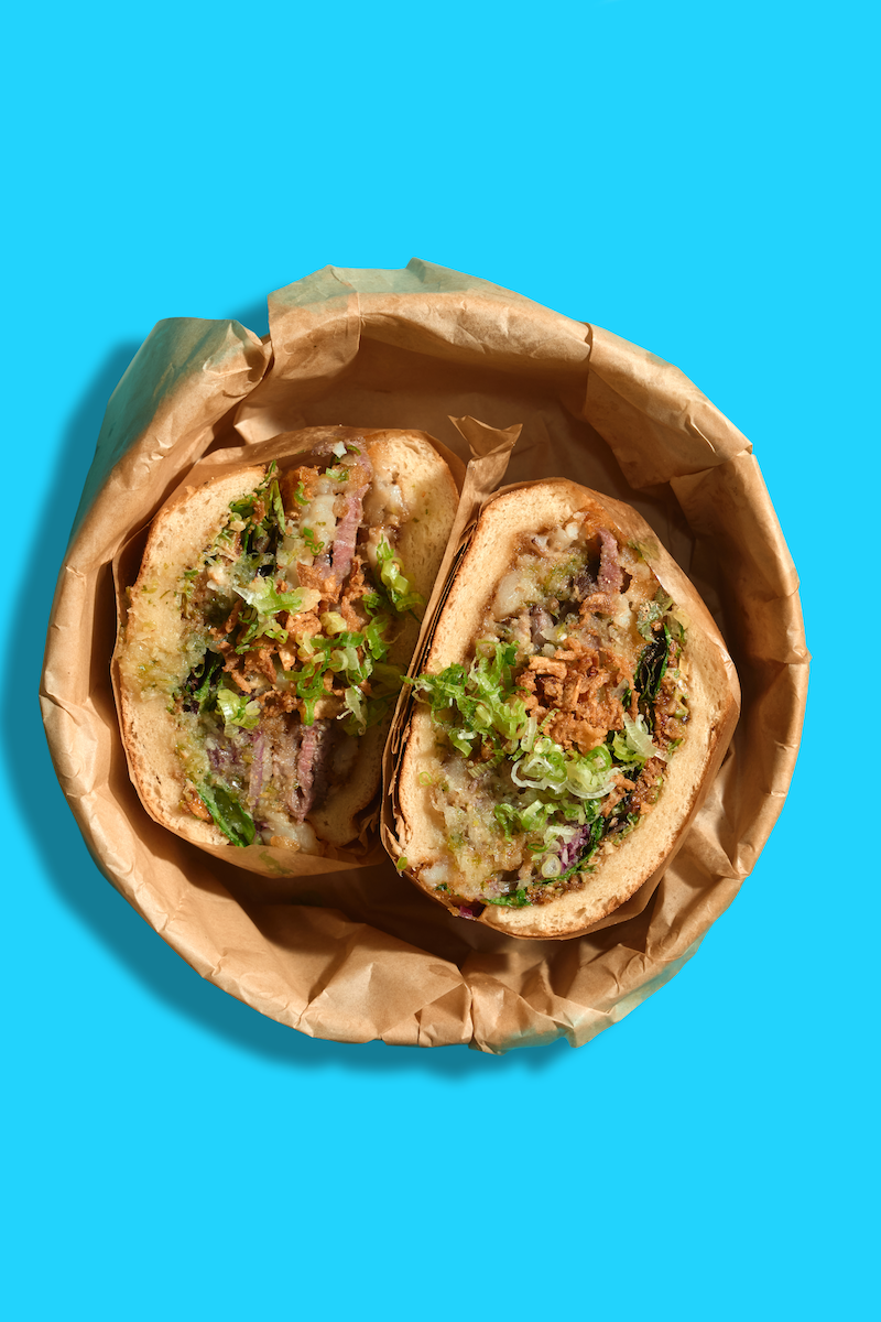 A sandwich cut in half, filled with meat, vegetables, and sauce, served in a brown paper lining.