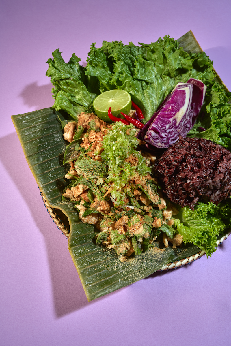 Traditional Thai dish served on a banana leaf with fresh lettuce, lime wedge, red chili peppers, purple eggplant, and a mound of black sticky rice.