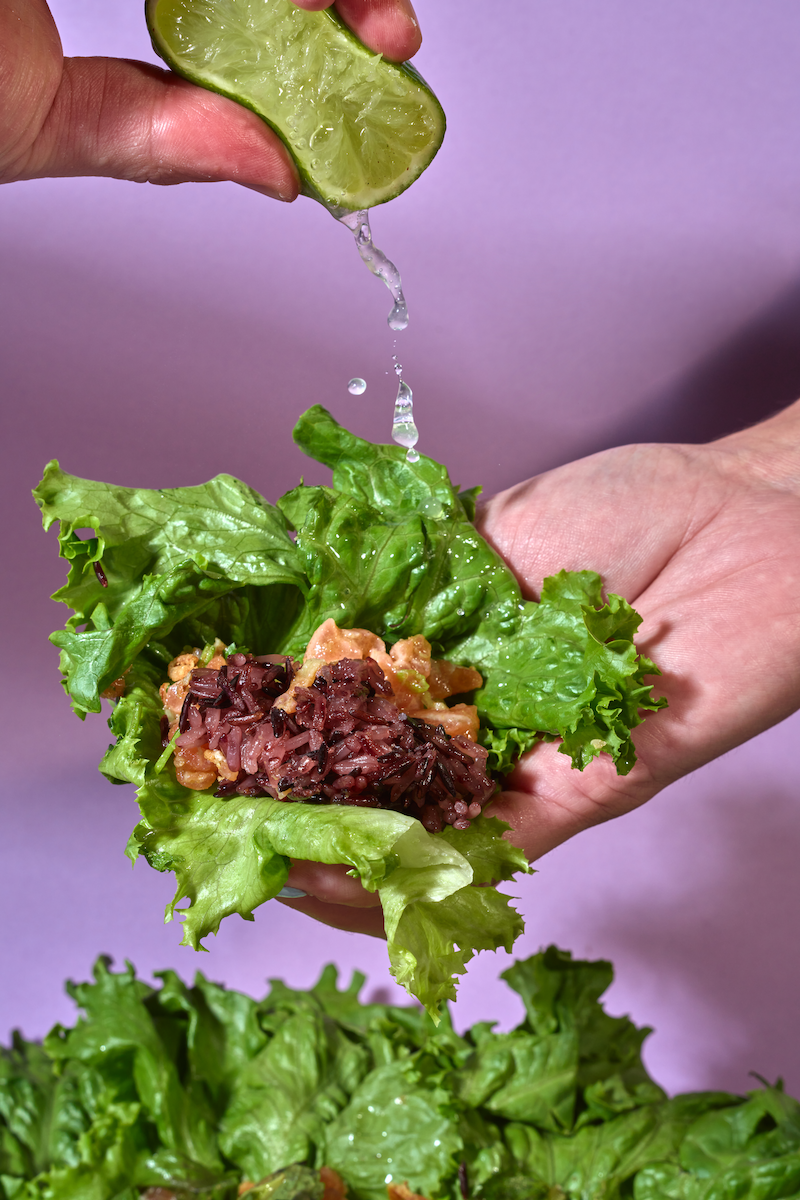 A hand holds a slice of lime above a lettuce wrap filled with rice and vegetables, with water droplets falling onto the wrap.