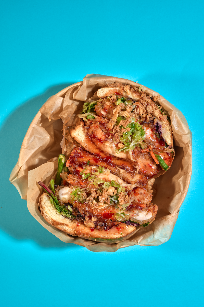 Close-up of a fried chicken sandwich with lettuce, green chili, red sauce, and crispy onion toppings inside a bun, in a brown paper wrapper on a blue background.