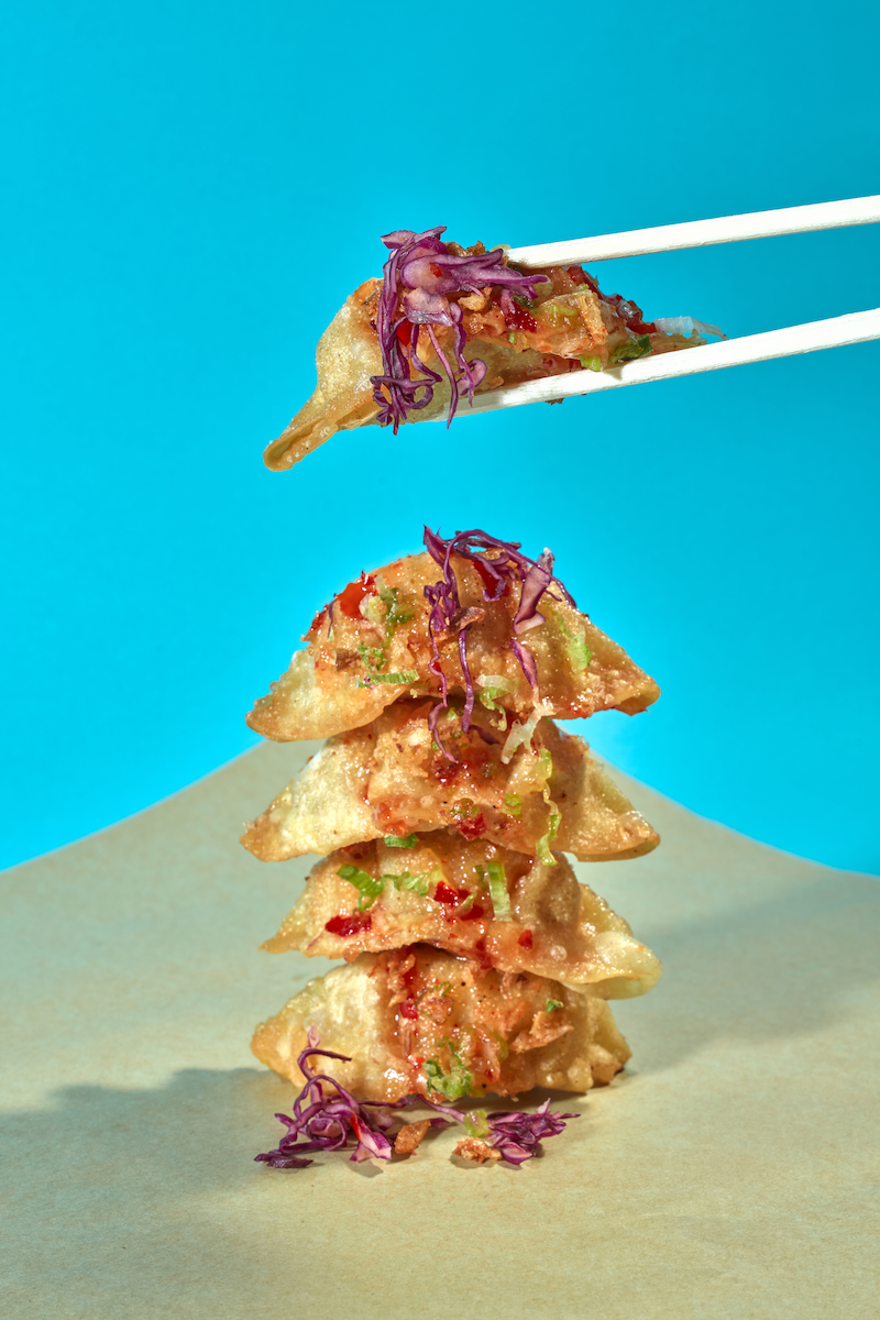 A stack of fried dumplings topped with chopped green onions and purple cabbage on a beige surface against a blue background, with some purple cabbage on the surface and chopsticks holding one dumpling.
