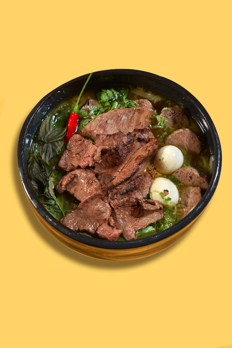 A bowl of Vietnamese pho containing beef slices, quail eggs, herbs, and a red chili pepper.