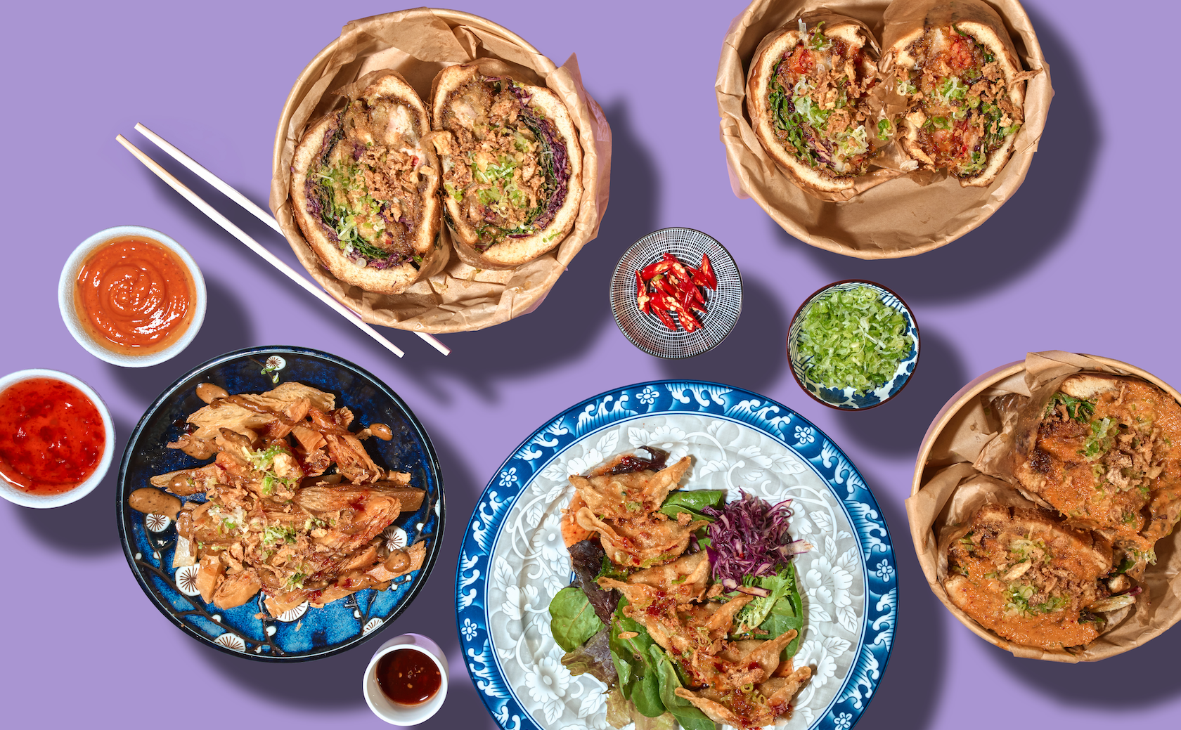 A top-down view of an assortment of Asian dishes on a purple surface, including three bowls of ramen with broth and toppings, a platter of fried vegetables, a salad with fried meat, and several small bowls of colorful sauces, garnishes, and condiments.
