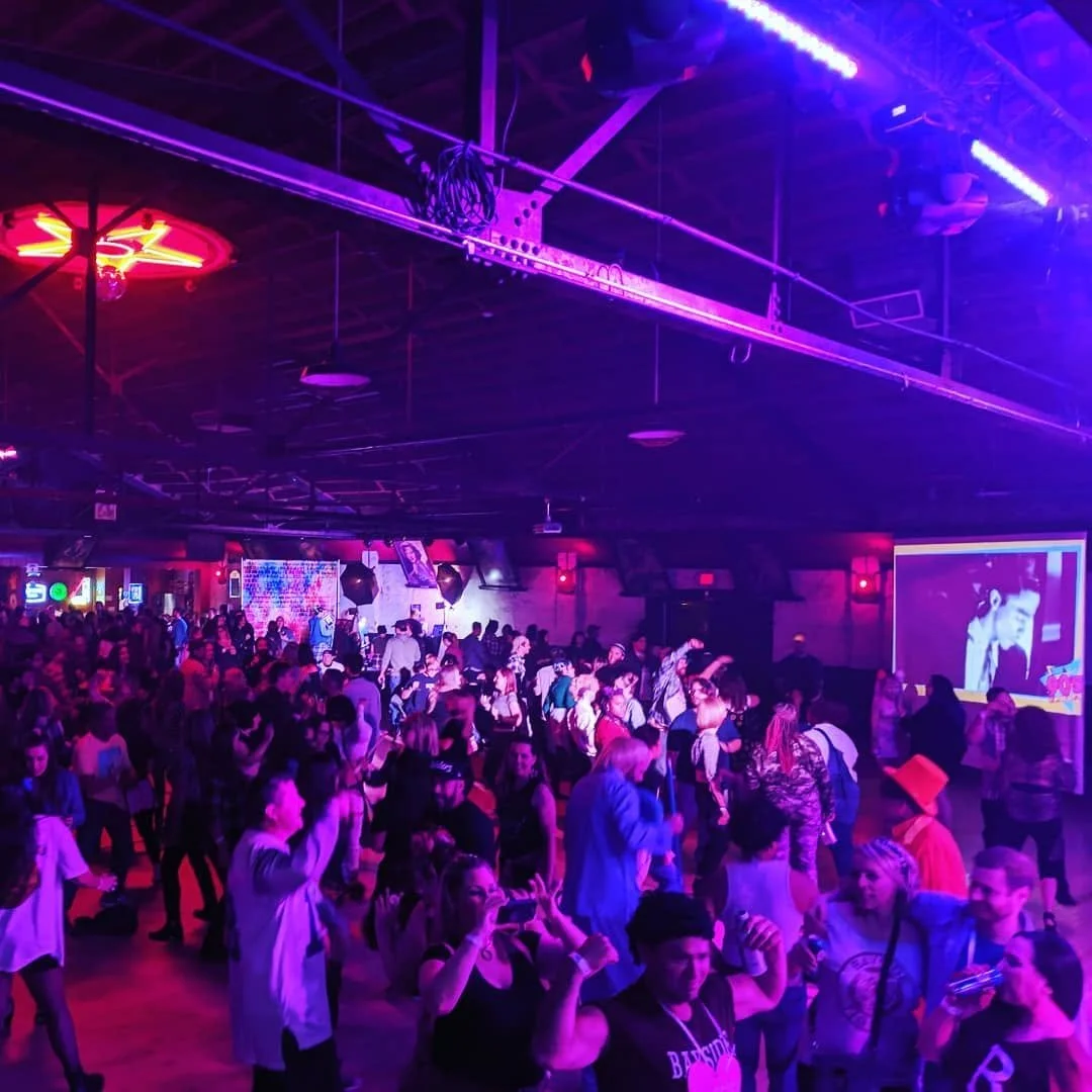 A crowded indoor dance club with purple and pink lighting, people dancing and socializing, and a large screen displaying visuals in the background.