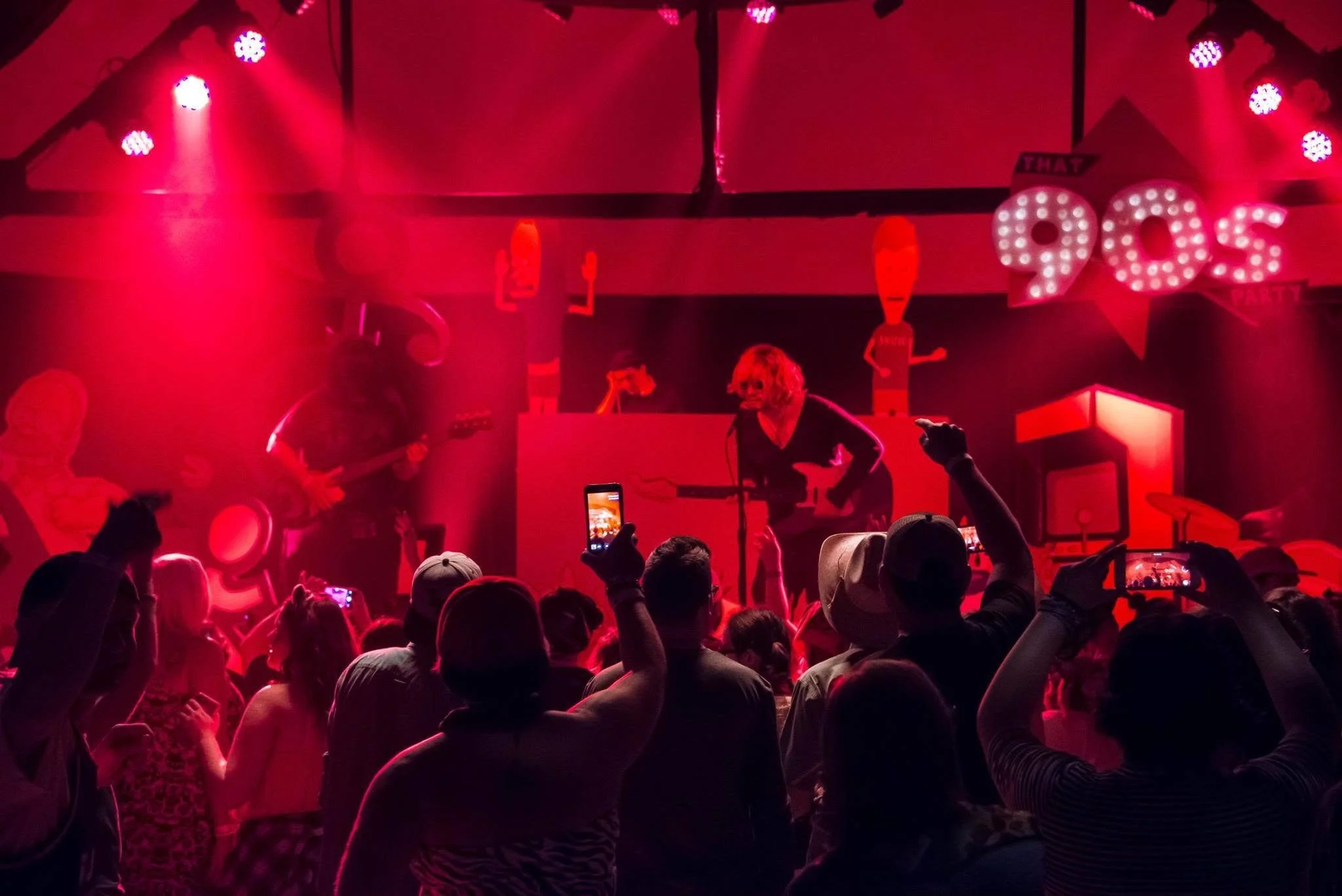 A lively indoor concert with a band performing on stage. The stage is lit with red and pink lights, and the crowd is raising hands and phones to capture the moment.