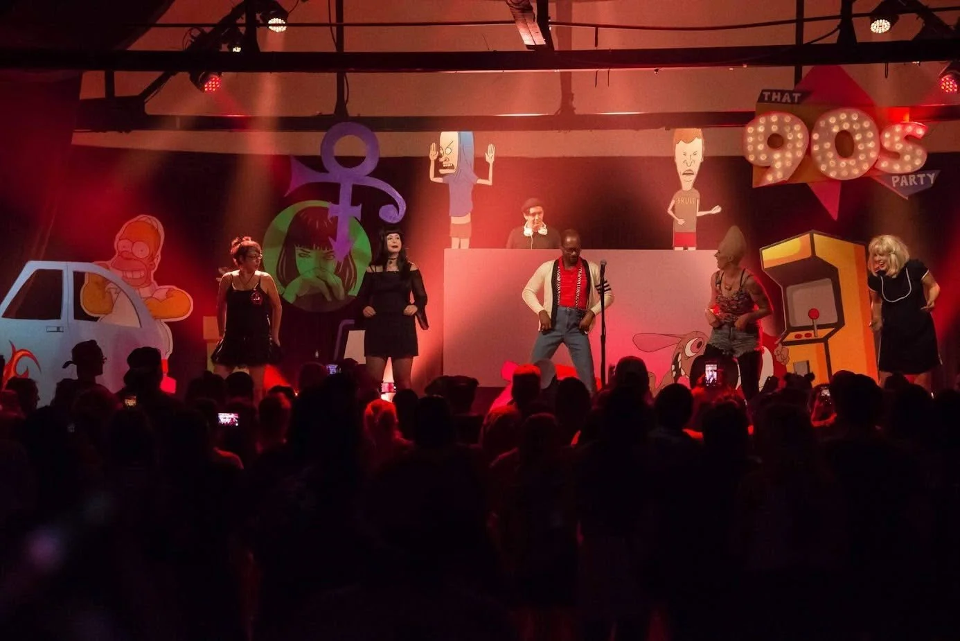 Actors performing a comedy or musical skit on stage at a '90s themed party, with colorful cartoon decorations and an audience watching.