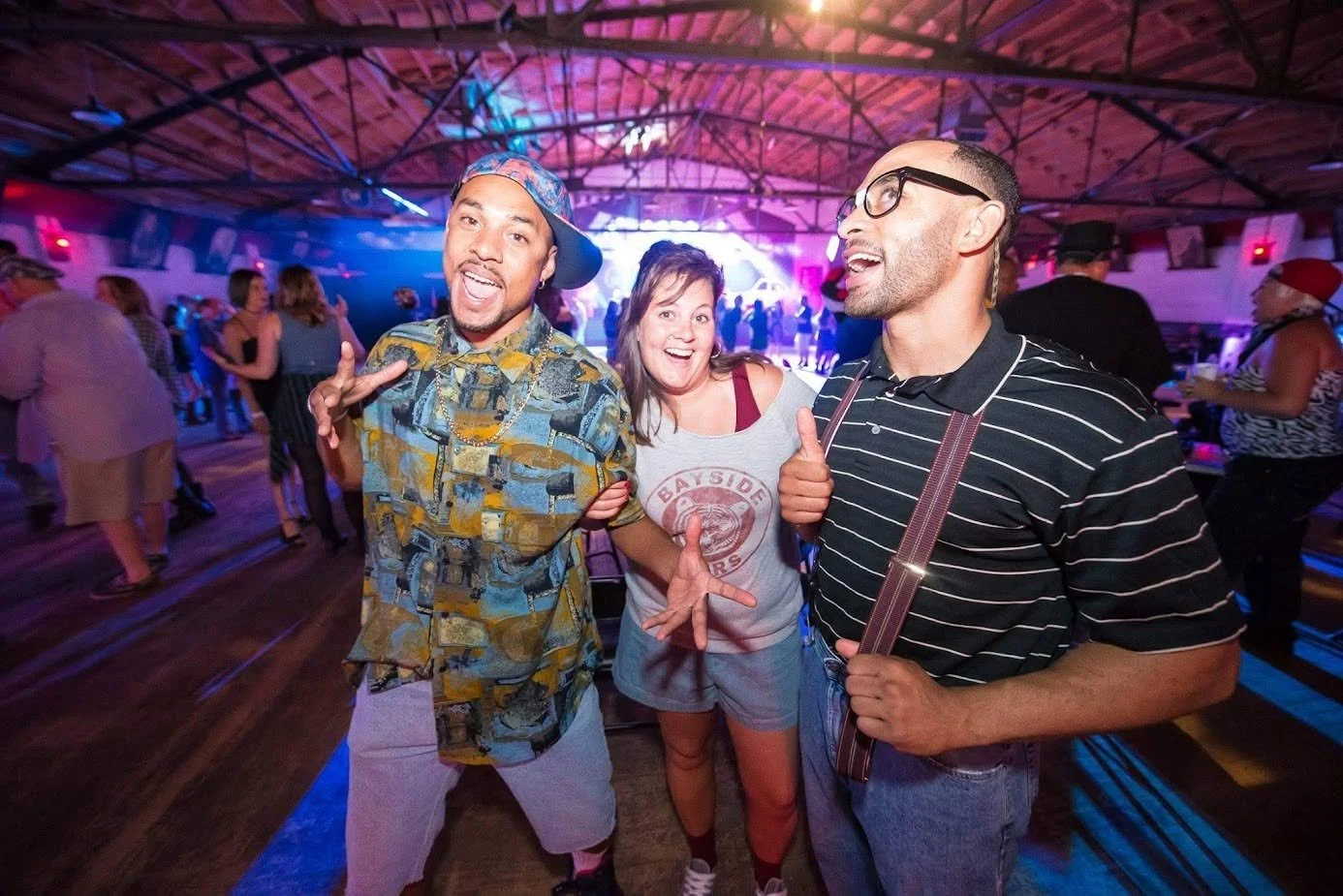 Three people smiling and posing for a photo at a lively indoor party or dance event with colorful lighting and other people dancing in the background.